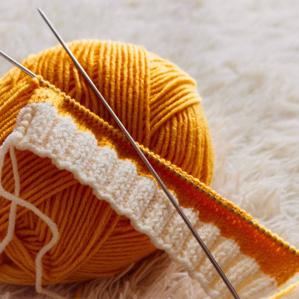Knitting needles working on a project with yellow yarn and a white textured border.