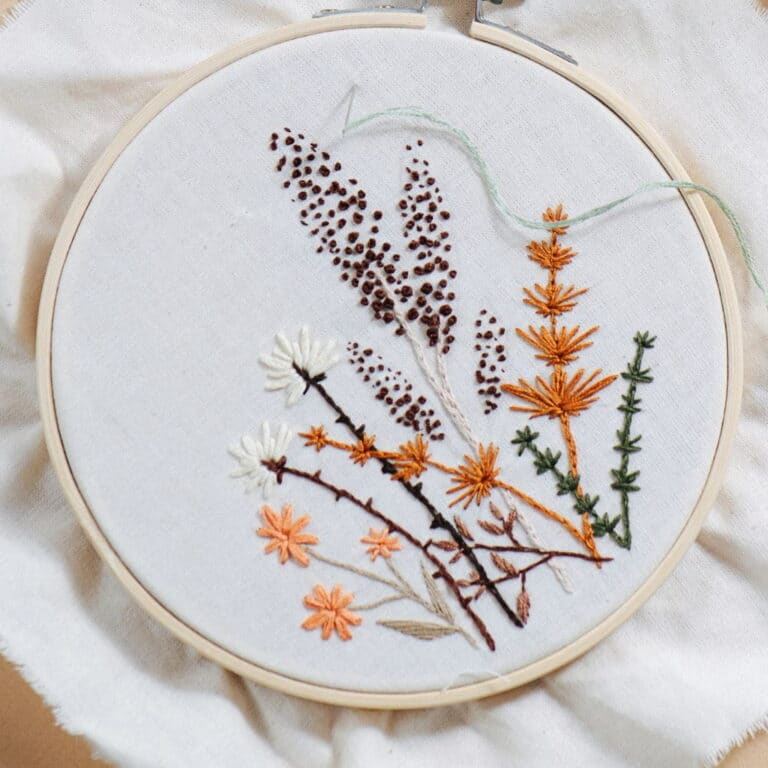 Embroidery hoop featuring a botanical design of wildflowers and grasses in shades of brown, orange, white, and green on white fabric.