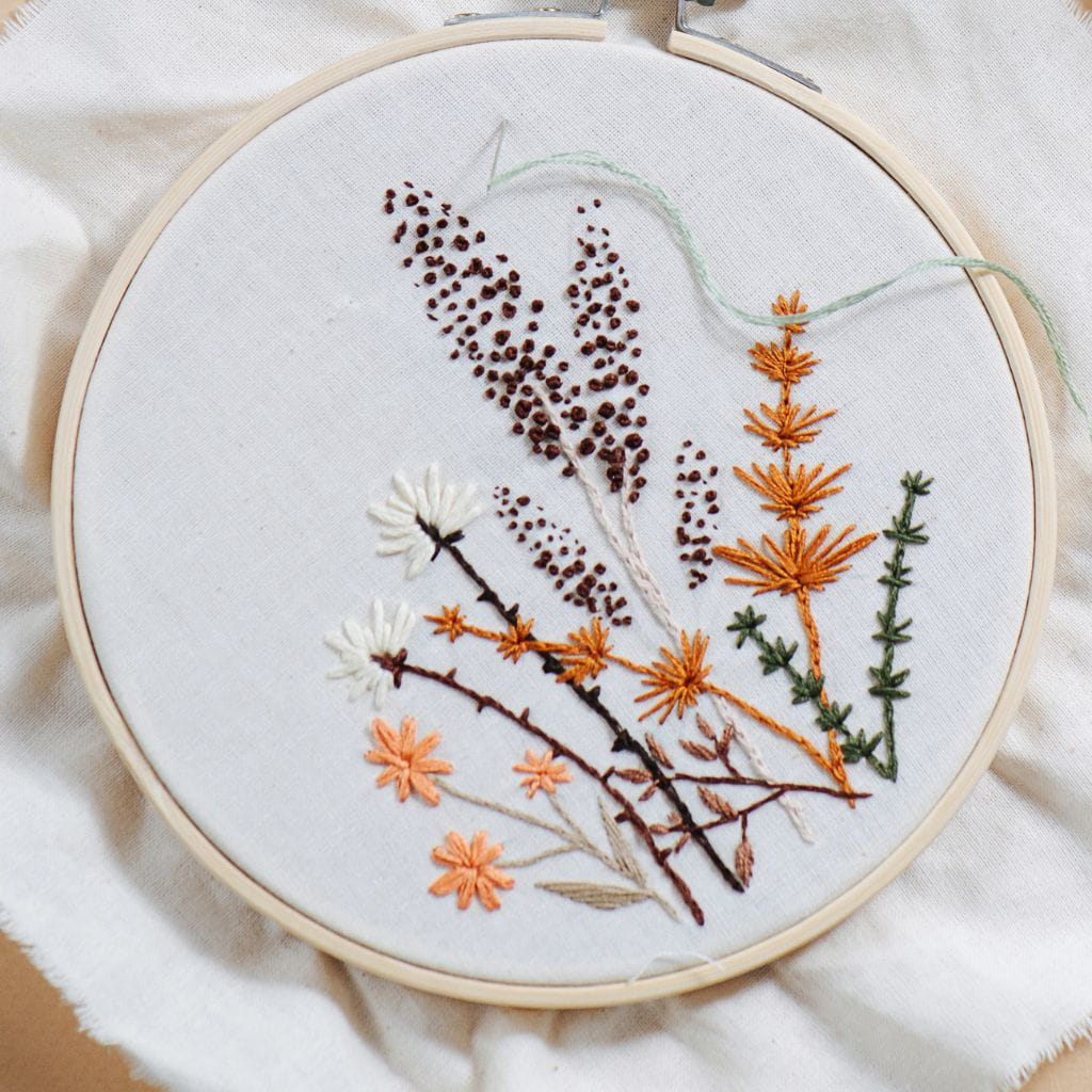 Embroidery hoop featuring a botanical design of wildflowers and grasses in shades of brown, orange, white, and green on white fabric.