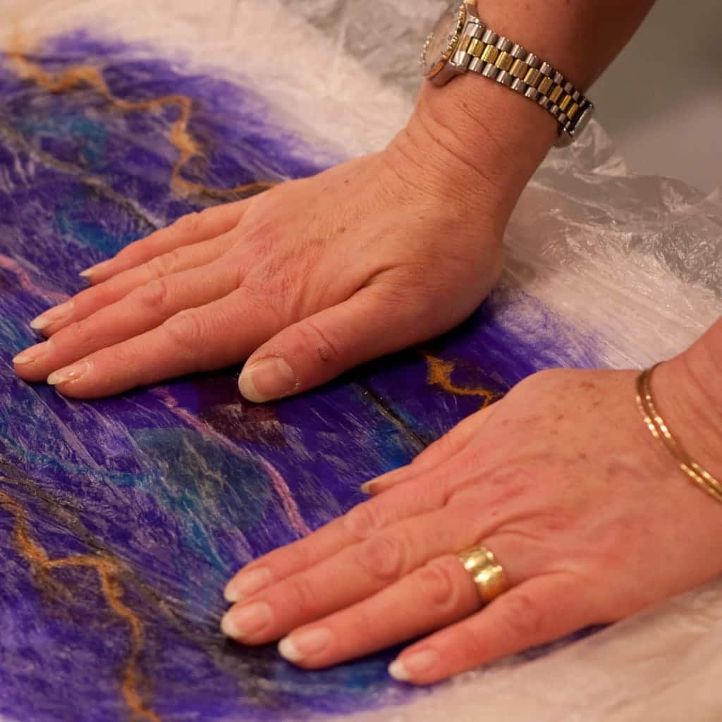 Hands pressing down on a piece of layered, blue and purple wool fiber art covered by a sheet of plastic.