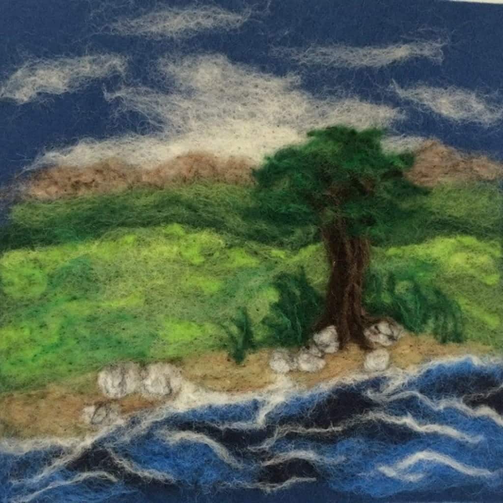 A needle-felted landscape featuring a single tree on a grassy shore next to a blue ocean, with distant hills and a cloudy sky.