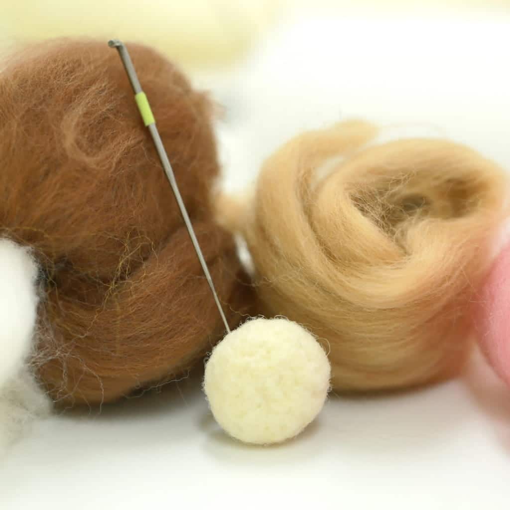 A needle felting needle inserted into a small white wool ball, positioned in front of mounds of unspun brown and tan wool.