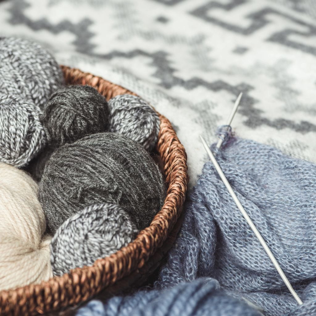 A woven basket filled with balls of grey and beige yarn, next to a piece of blue knitted fabric on knitting needles.