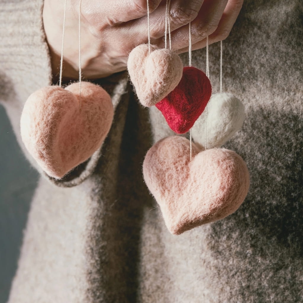 Hands hold a cluster of five hanging felt hearts in shades of pink, red, and white.