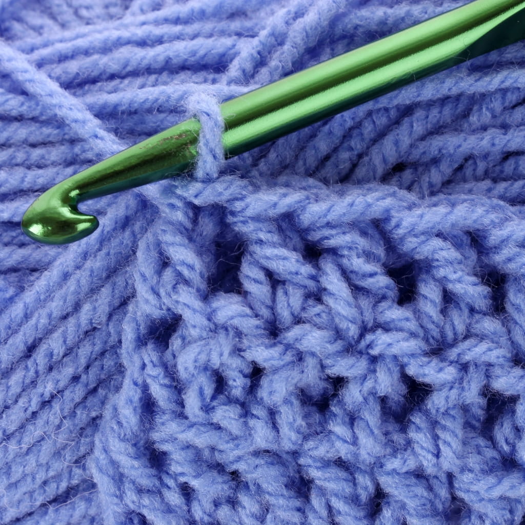 A green crochet hook pulling a loop of periwinkle yarn through a stitch in a piece of crocheted fabric.