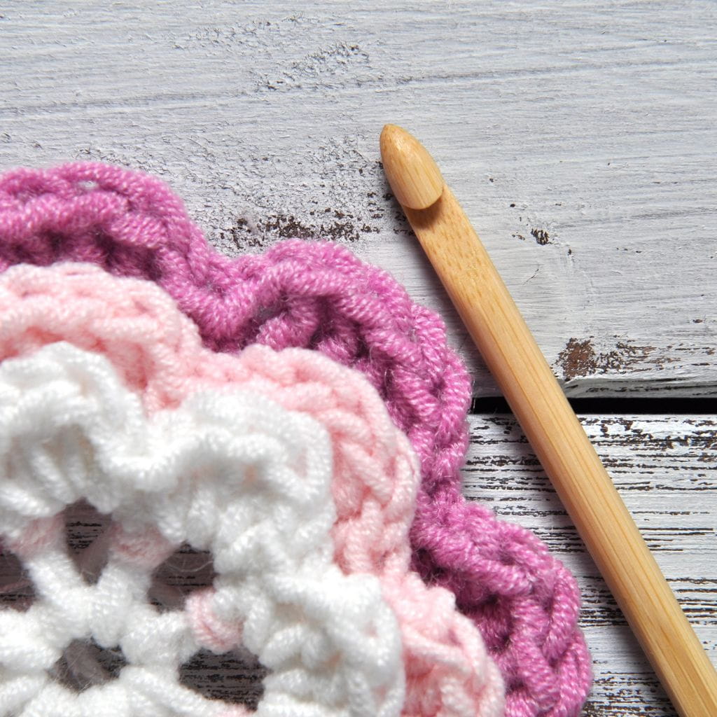 A wooden crochet hook resting next to a white, light pink, and dark pink crocheted doily on a white wooden surface.
