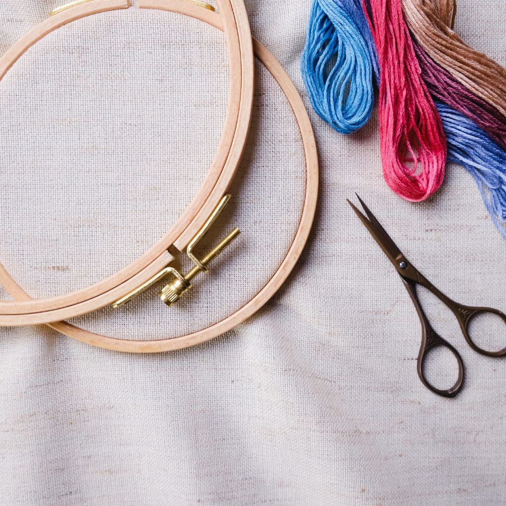 Embroidery for beginners supplies. A wooden embroidery hoop, bundles of colorful embroidery floss, and small scissors on a piece of fabric.