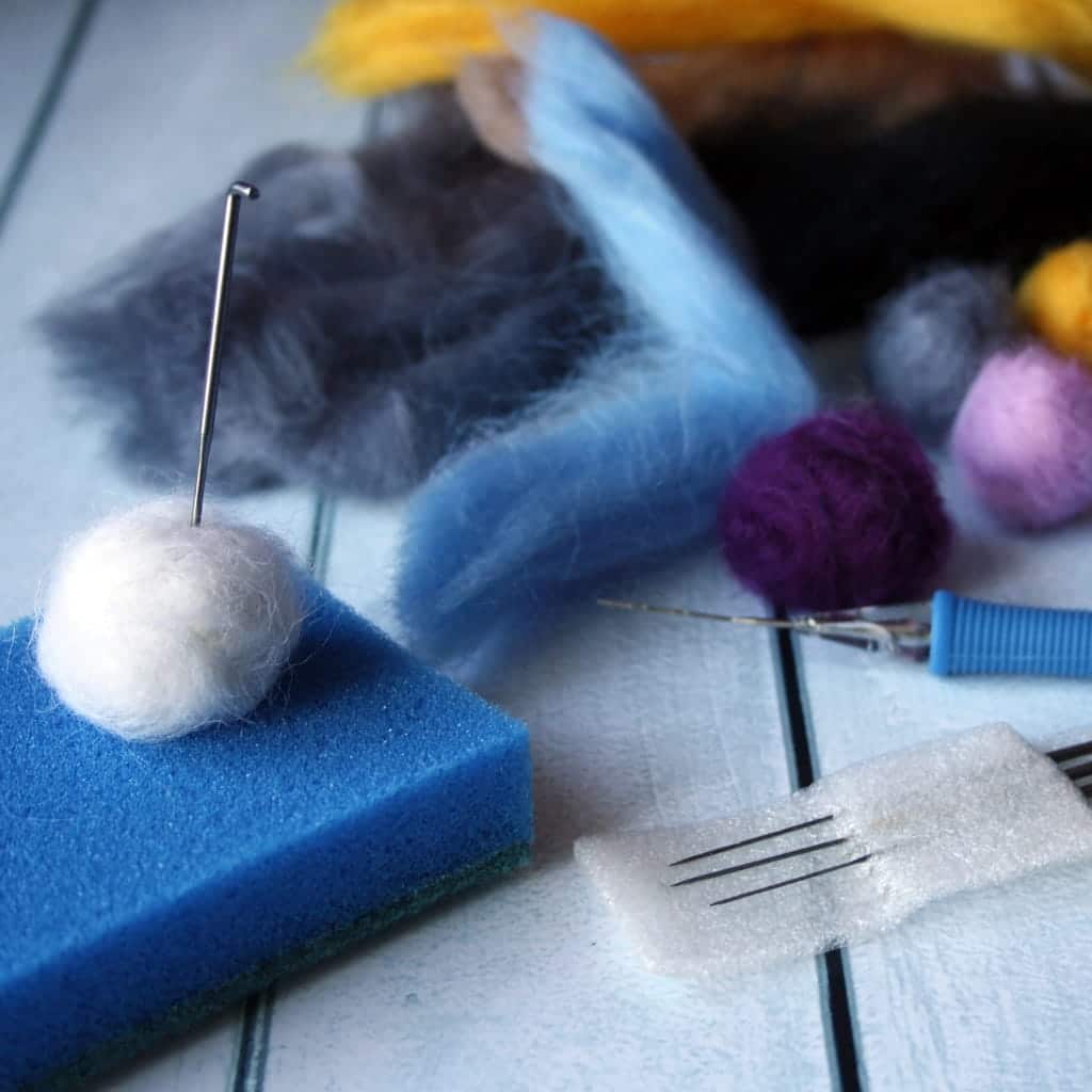 Needle felting supplies including colorful wool roving, a blue foam pad with a white wool ball, and felting needles.