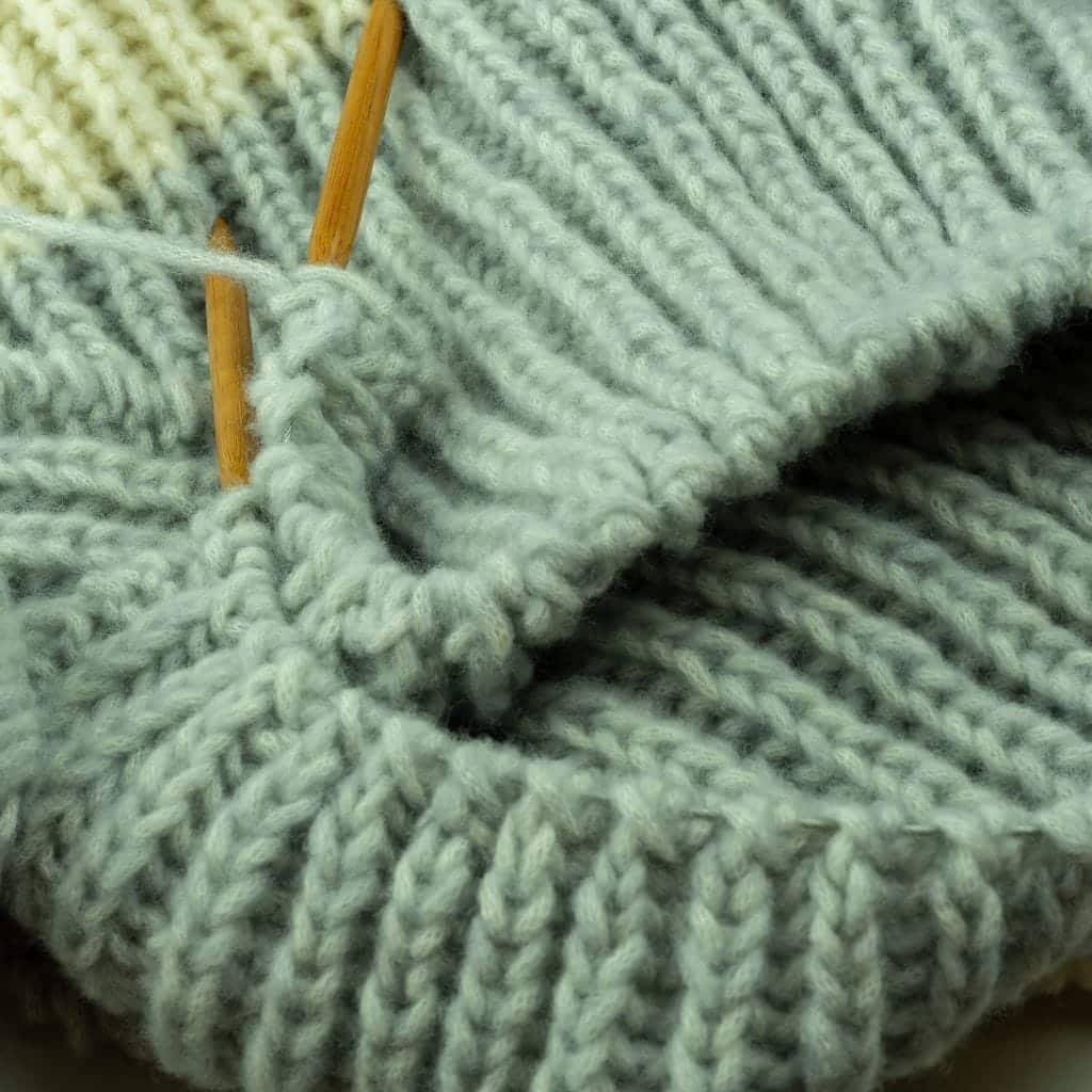 Knitting projects for beginners. Close-up of a ribbed knit project on wooden knitting needles, transitioning from light green to off-white yarn.